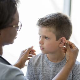 A little boy is sitting on an examining table in a doctors office - a healthcare professional is implanting a hearing aid for the child.