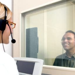 Man getting a hearing test in a hearing booth with an audiologist on the other side of the booth's glass