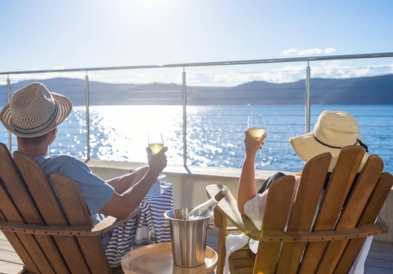 A couple drinks wine on deck chairs on a cruise ship deck overlooking the water